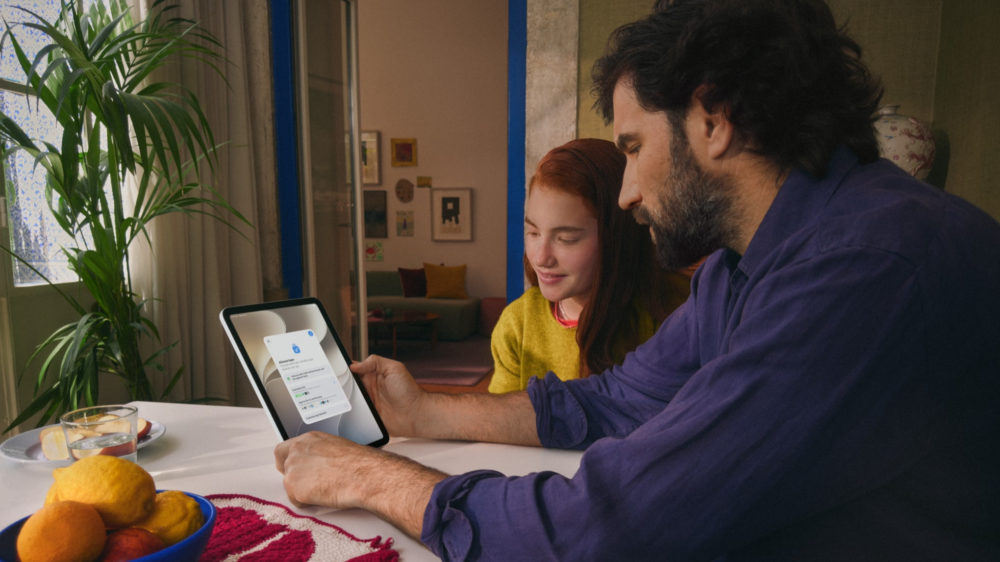 Padre e hija usando tableta en sala moderna, aprendiendo juntos. Frutas y vaso en la mesa completan la escena acogedora.