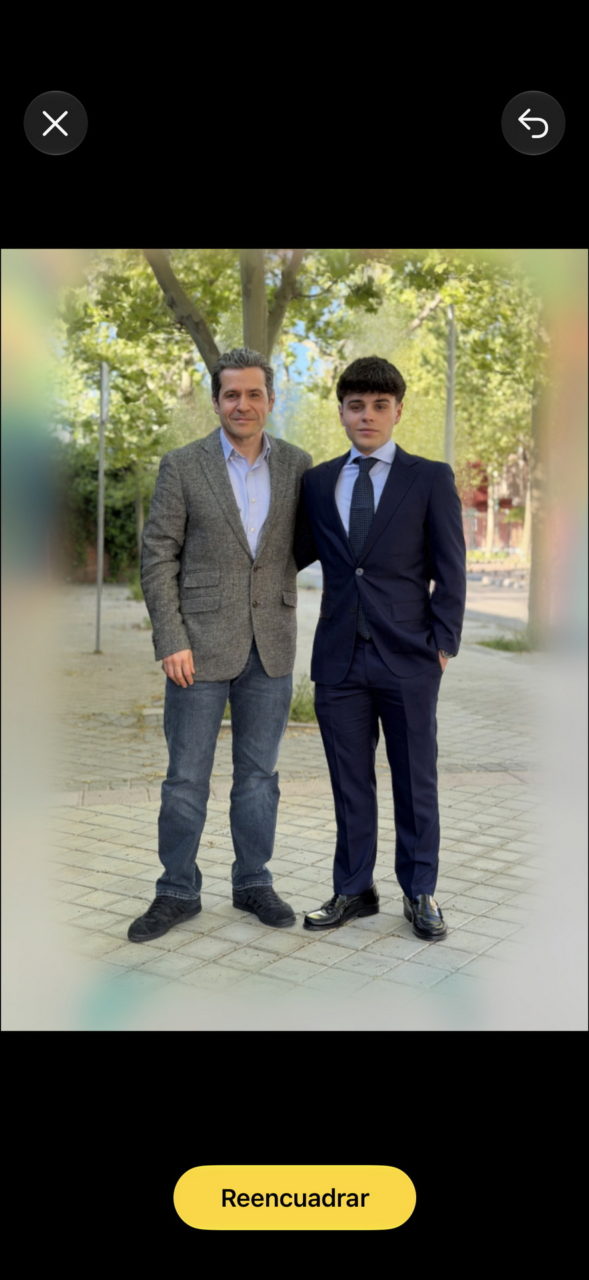 Un hombre y un joven con traje posan en un parque. Evento formal al aire libre con árboles de fondo.