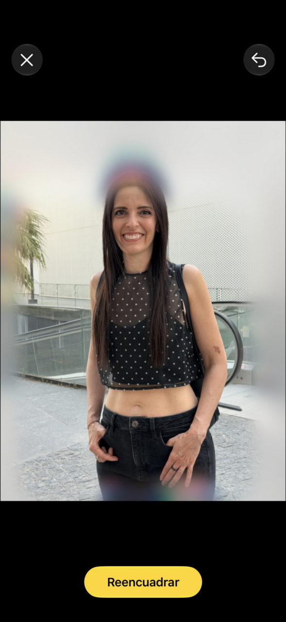 Mujer sonríe con top de lunares y jeans negros frente a edificio moderno con plantas.