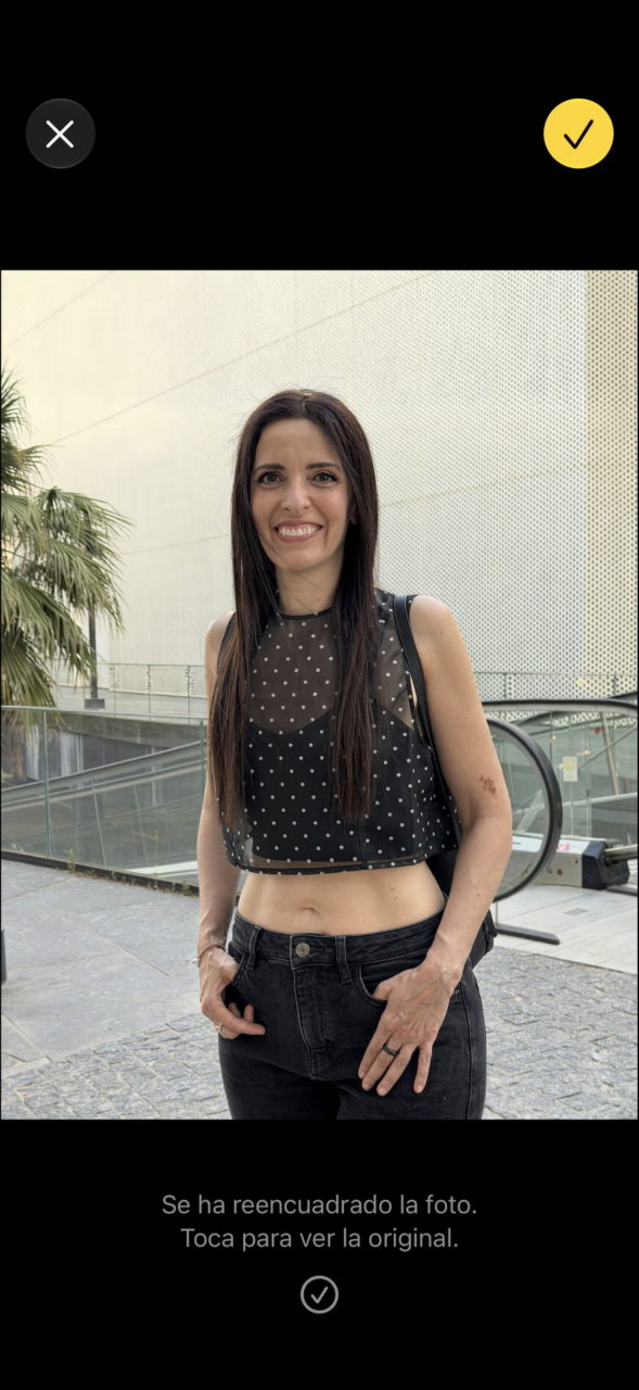 Joven sonriendo con top negro y pantalón oscuro, fondo urbano con plantas y paredes modernas.