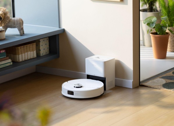 Robot aspirador situado en una sala luminosa, con plantas y muebles modernos, cargando en su base.