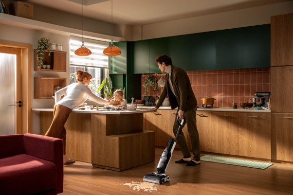 Familia limpiando cocina moderna, padre con aspiradora, madre con bebé en encimera, ambiente acogedor y luminoso.