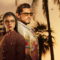 Dos personajes con gafas de sol al atardecer, rodeados de palmeras. Ambiente tropical y relajado.