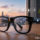 Gafas inteligentes en una mesa con íconos holográficos de navegación, cámara y música; vista de ciudad al atardecer.