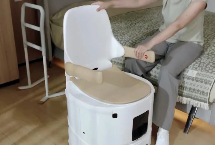 Persona ajustando una silla de inodoro portátil blanca junto a una cama, ideal para personas con movilidad reducida.