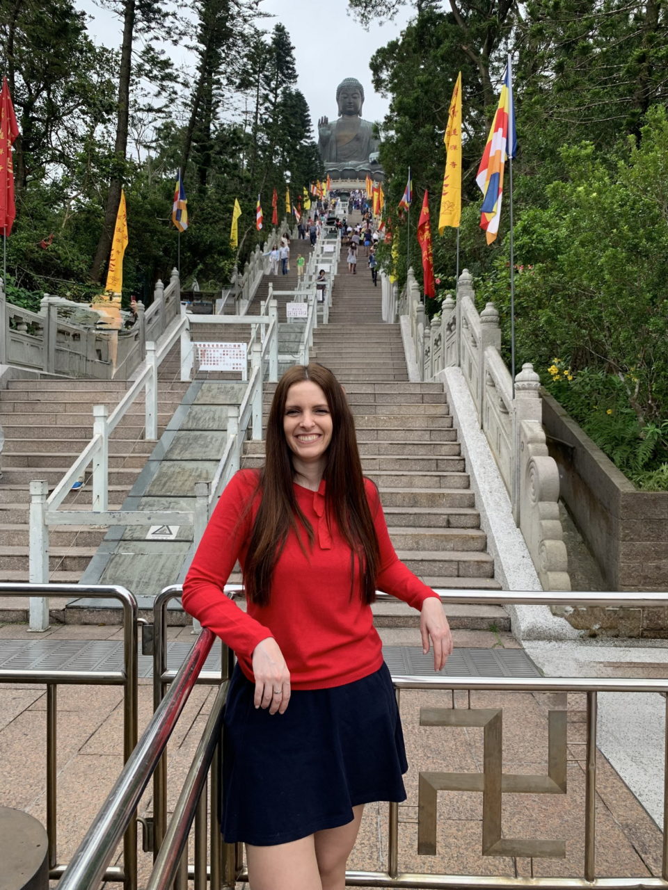 Mujer en ropa roja sonriendo frente a la escalera al Gran Buda en Hong Kong, rodeada de banderas y vegetación.