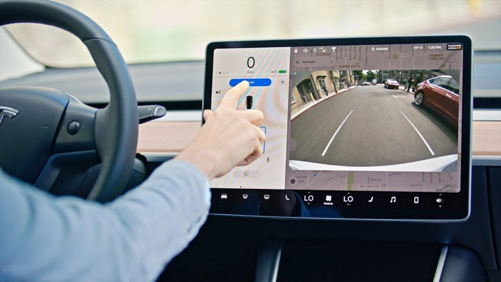 Tesla prepara una gran mejora para que sus coches aparquen donde tú quieres Conductor interactuando con pantalla táctil de auto eléctrico mostrando cámara trasera y controles de navegación.