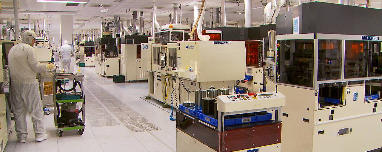 Fábrica de semiconductores con máquinas avanzadas y trabajadores en trajes protectores en sala limpia.