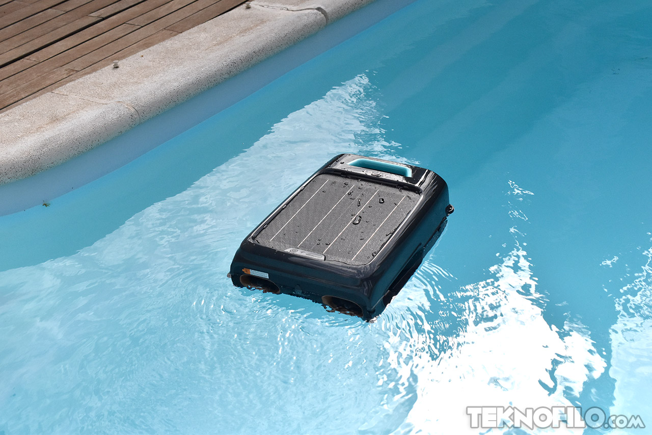 Robot limpiador de piscinas flotando en agua azul clara junto al borde de madera.
