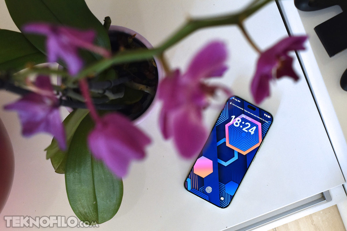 Smartphone con pantalla geométrica junto a una orquídea morada sobre mesa blanca. Tecnología y naturaleza.