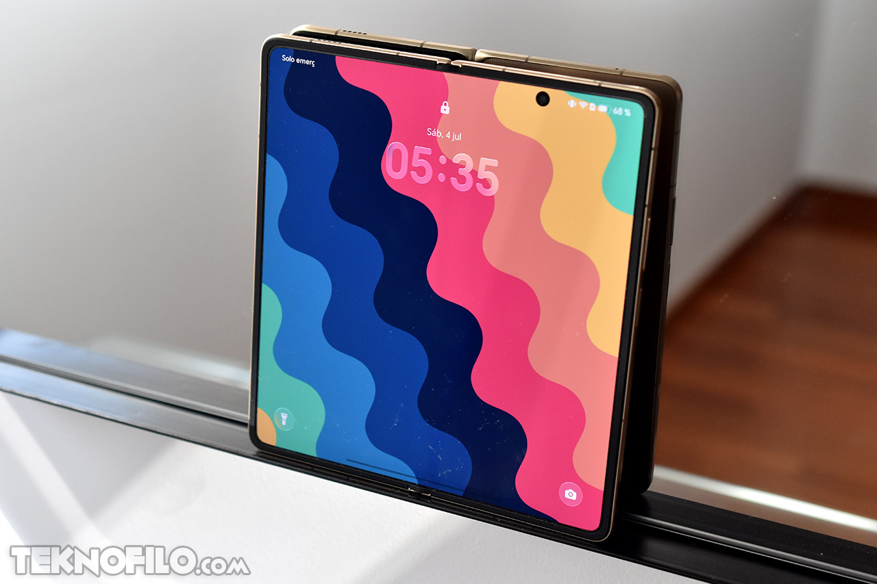 Smartphone plegable con pantalla colorida en un soporte de vidrio, mostrando la hora 05:35 y fondo de ondas multicolores.