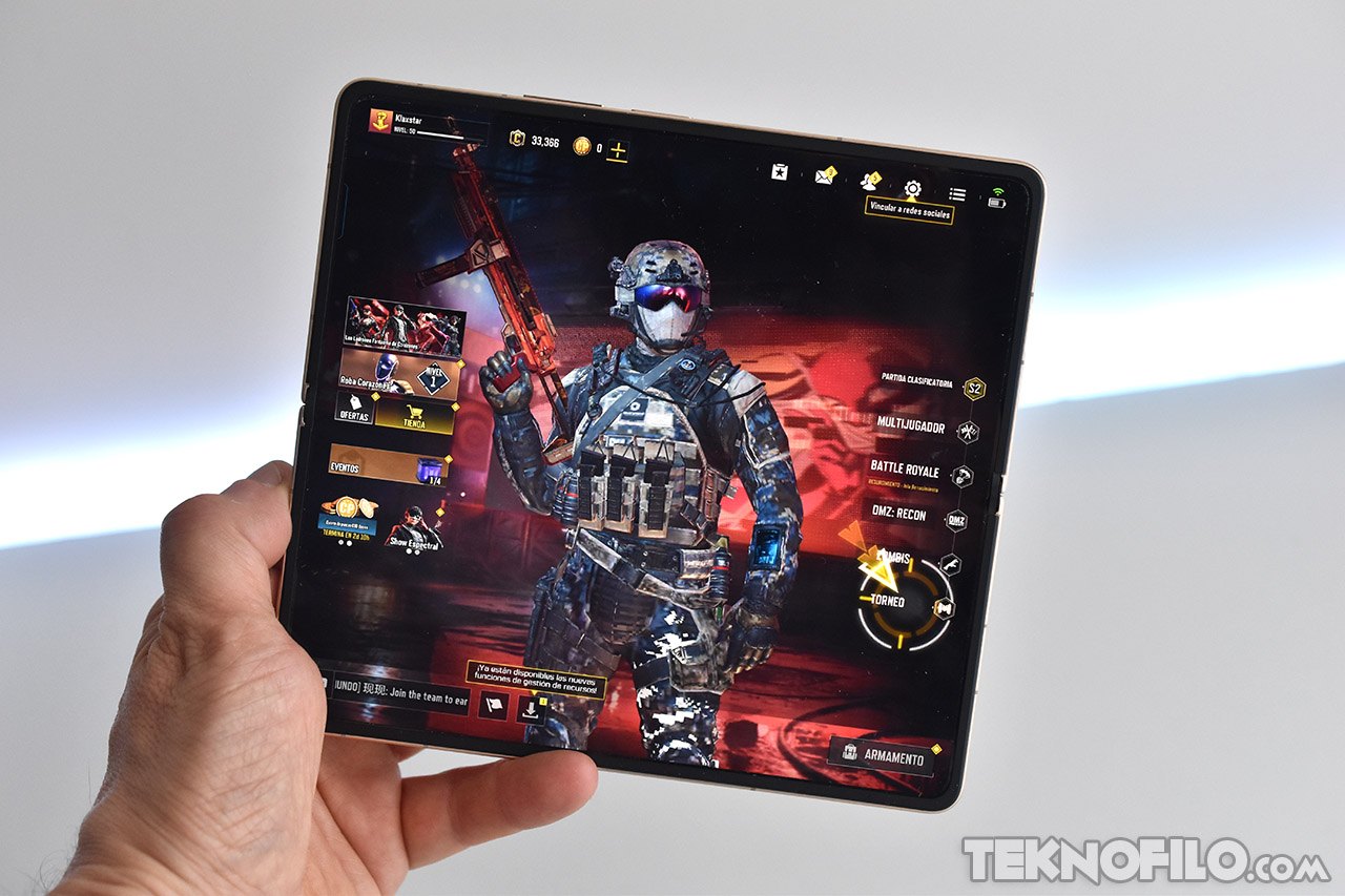 Jugador con traje táctico en pantalla de juego móvil en tableta plegable, demostrando características del juego.