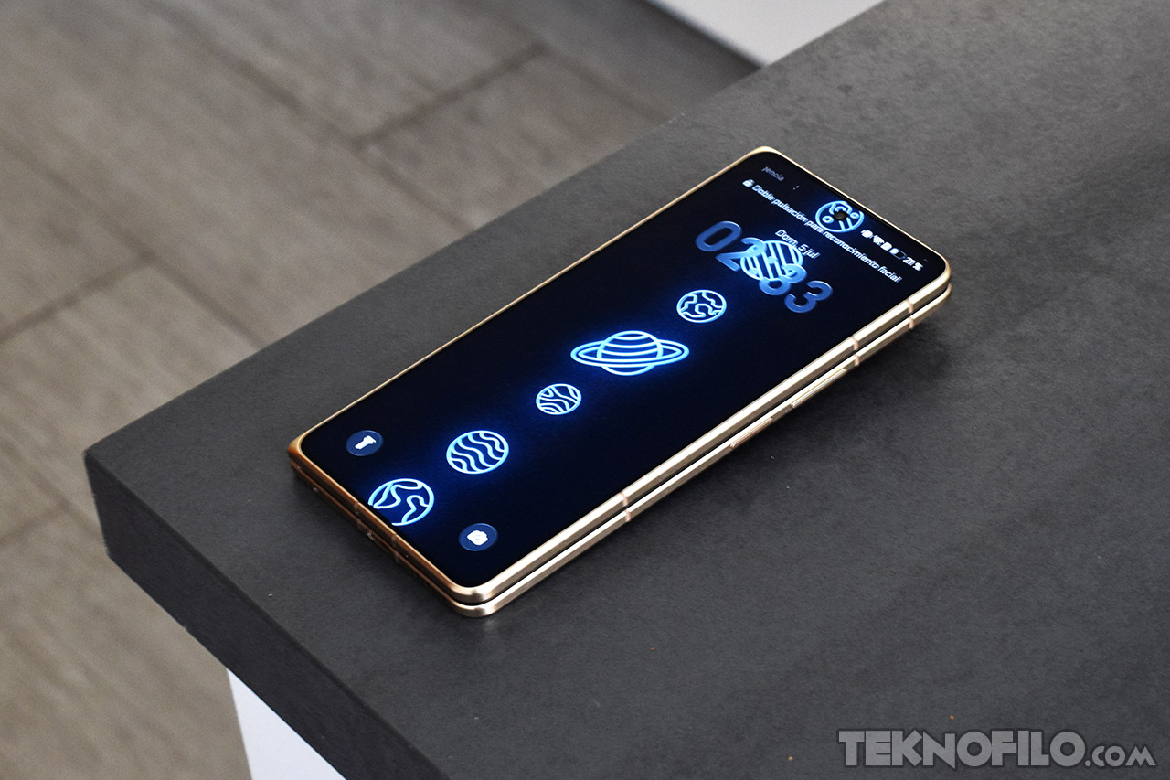 Smartphone plegable dorado con pantalla encendida mostrando diseño de planetas en una mesa gris moderna.