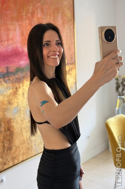 Mujer tomando selfie con móvil dorado en una habitación colorida, arte de fondo y sonrisa radiante.