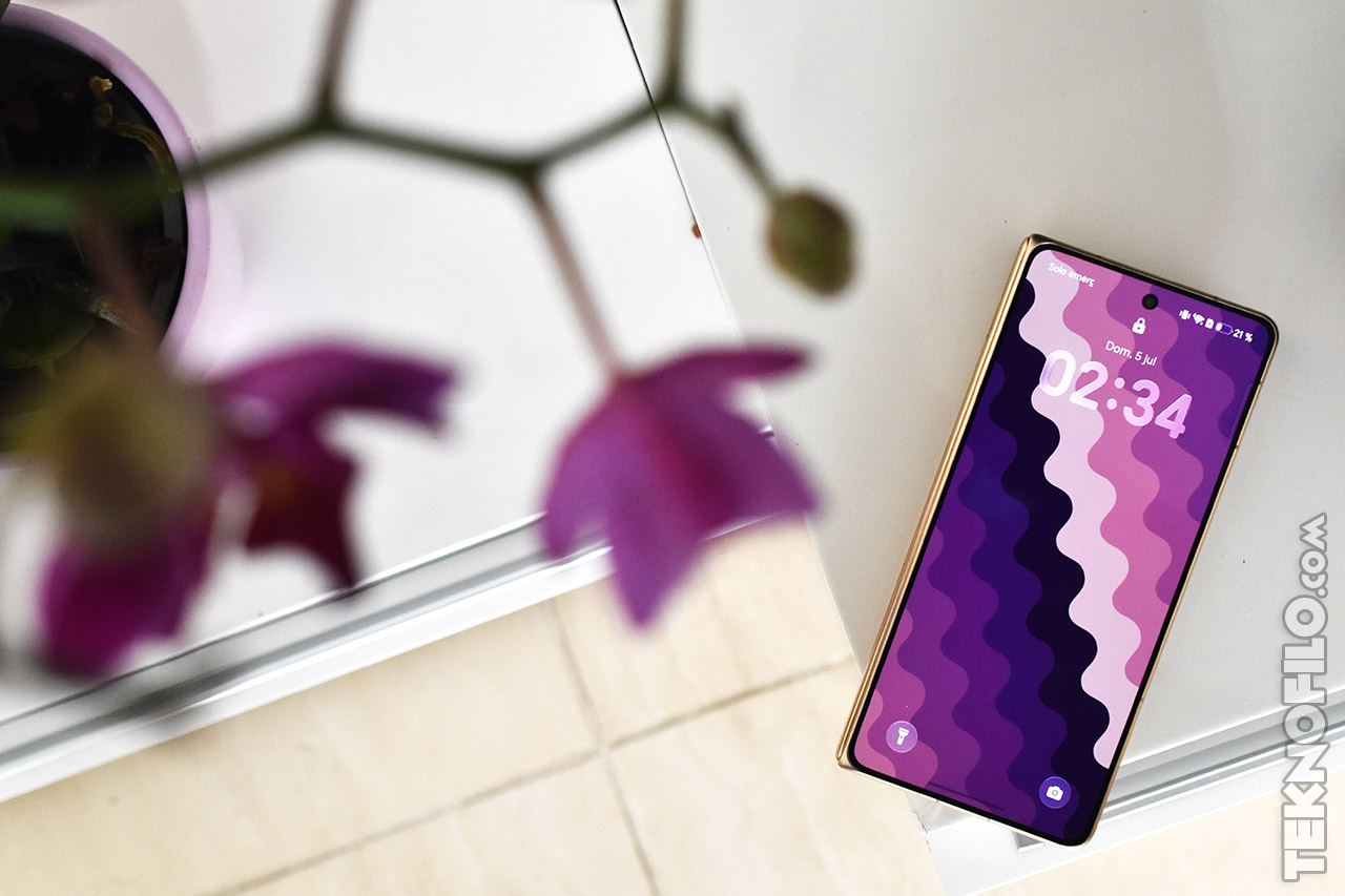 Teléfono inteligente con pantalla de fondo colorida sobre mesa blanca junto a una orquídea morada.