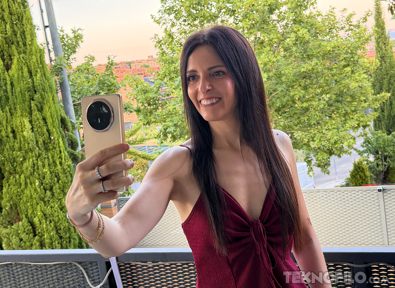 Mujer tomándose un selfie con un teléfono inteligente en un balcón, rodeada de vegetación.