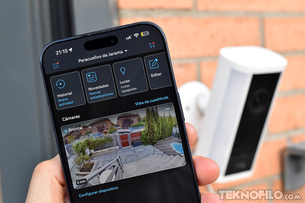 Smartphone mostrando aplicación de cámara de seguridad doméstica frente a un timbre conectado en una pared.