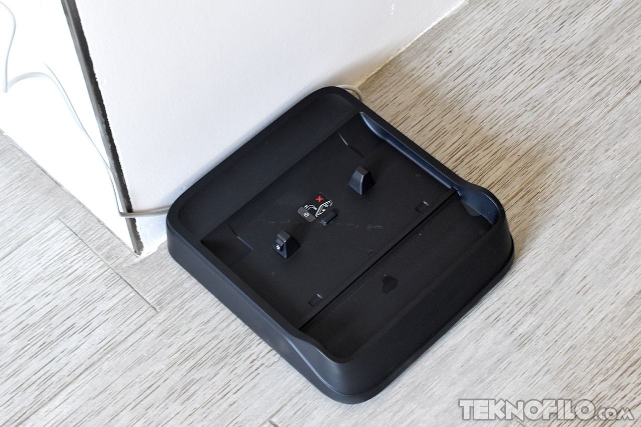 Base de carga para robot aspirador ubicada en el suelo junto a una pared blanca.