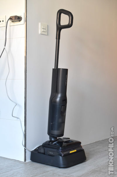 Aspiradora en estación de carga, conectada a la pared, sobre suelo de madera clara.