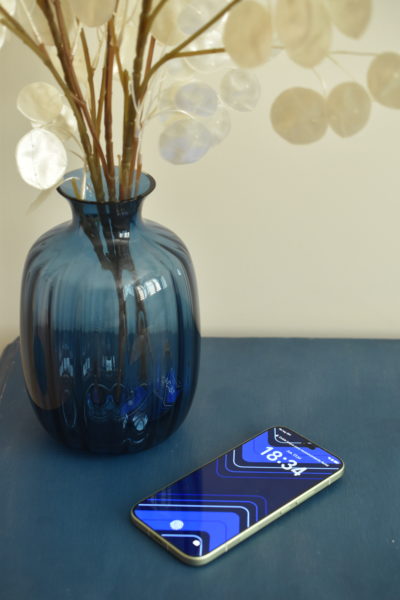 Teléfono móvil moderno sobre mesa azul junto a jarrón de cristal azul con flores decorativas.