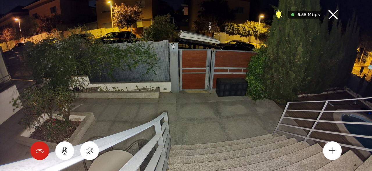 Patio exterior con escalera y puerta cerrada bajo iluminación nocturna, rodeado de plantas y arbustos.