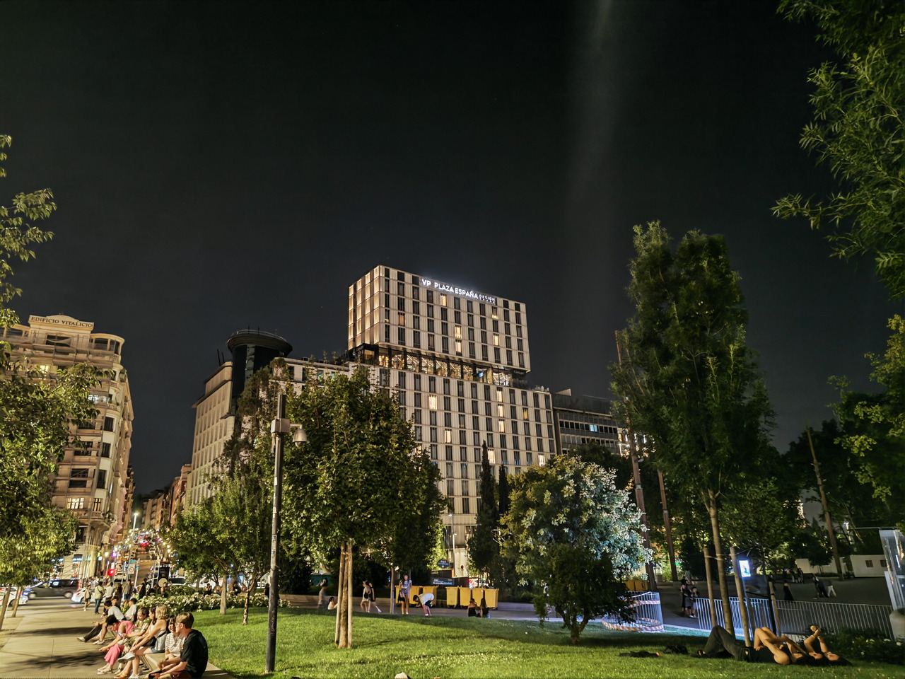 Edificio iluminado en la noche con personas descansando en un parque cercano, rodeado de árboles.