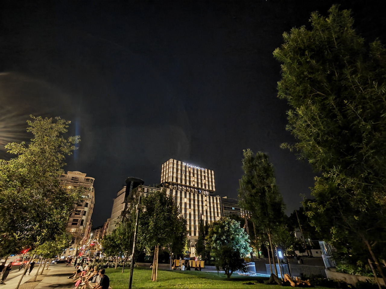 Vista nocturna de un parque urbano con edificios iluminados y gente paseando en una ciudad moderna.