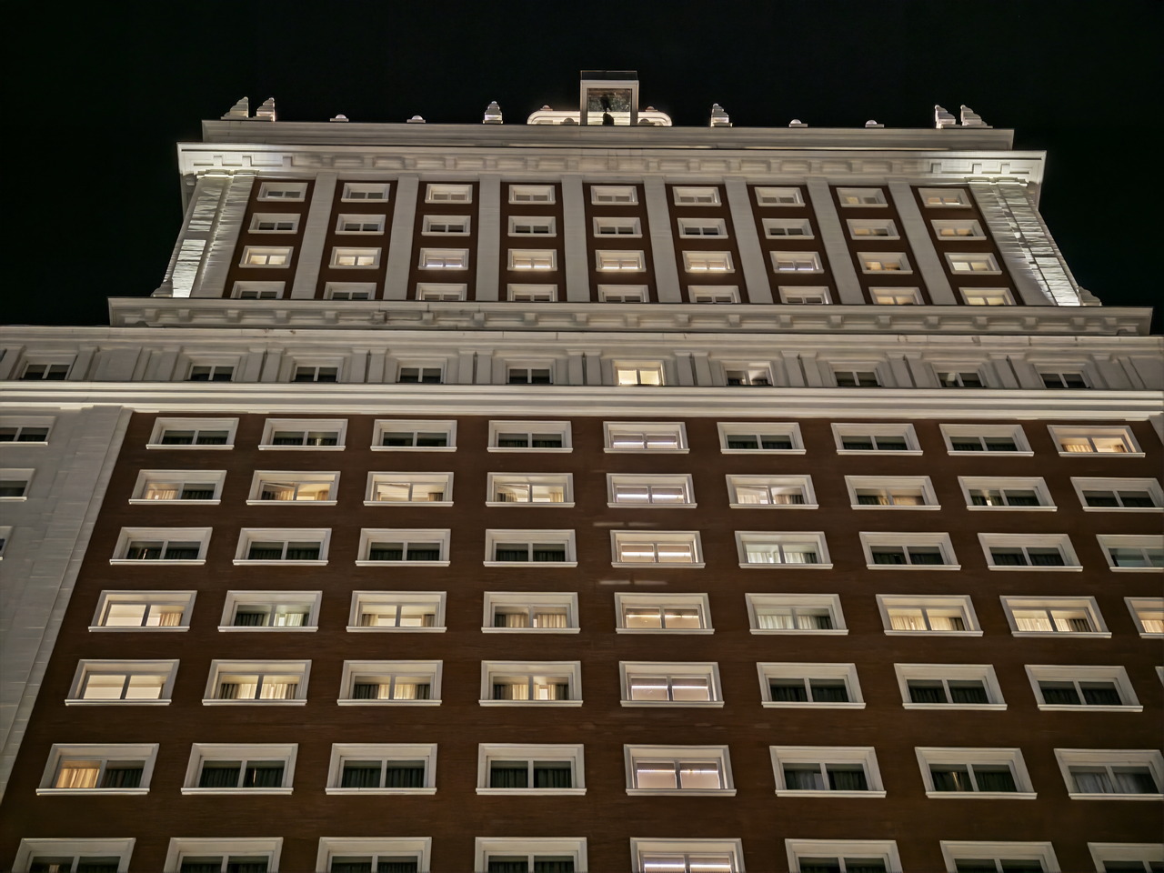 Edificio iluminado por la noche con ventanas encendidas, visto desde abajo, resaltando su fachada clásica.
