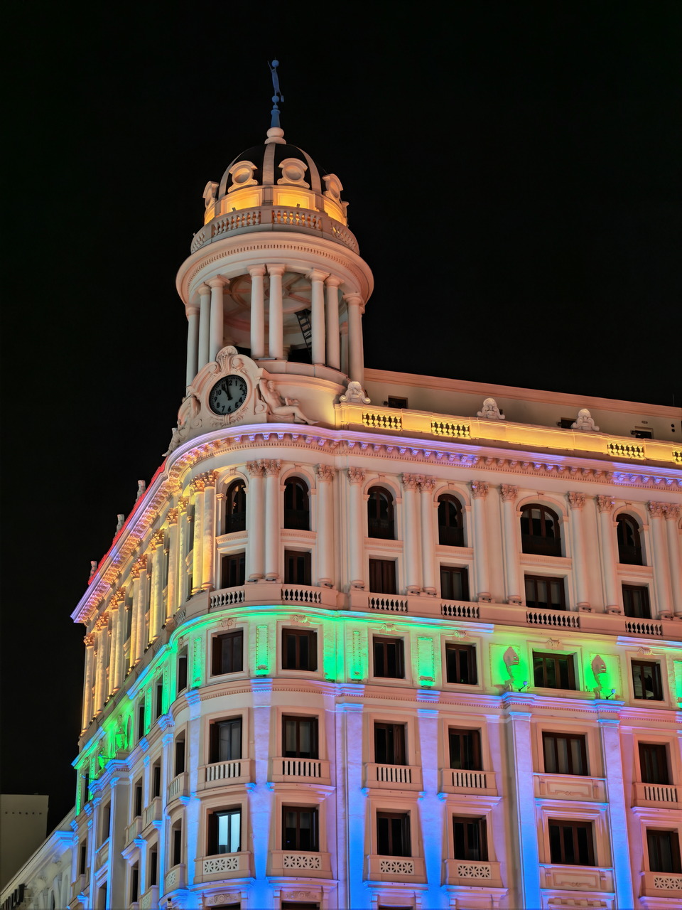 Edificio histórico iluminado con luces de colores en la noche, resaltando su arquitectura y detalles neoclásicos.