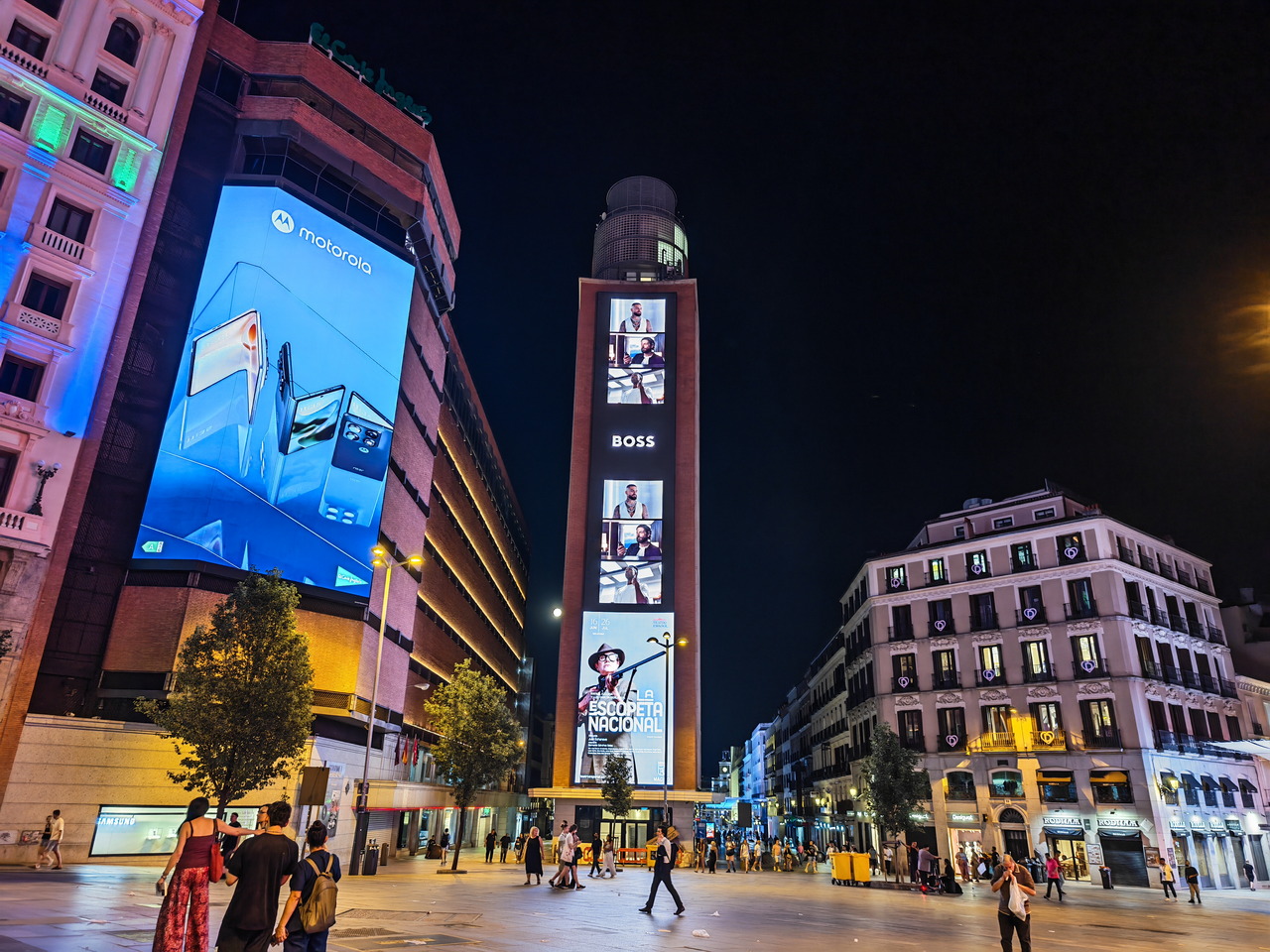 Carteles publicitarios iluminan la noche en una concurrida plaza urbana, destacando marcas tecnológicas y de moda.