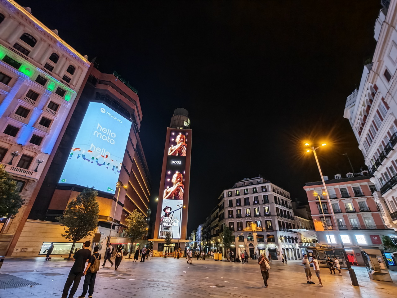 Plaza nocturna en Madrid con carteles publicitarios iluminados y gente paseando.