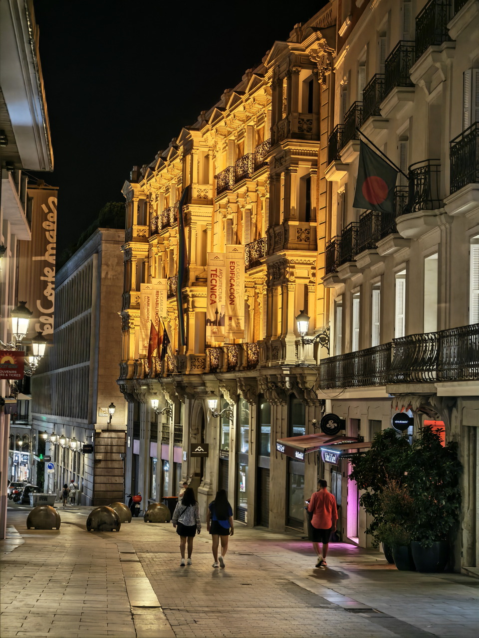 Calle iluminada de noche en el centro de la ciudad, con personas caminando y edificios históricos al fondo.