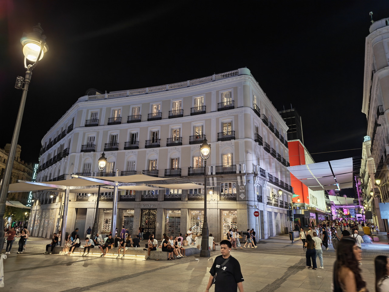 Calle concurrida en una ciudad europea de noche, con edificio iluminado y personas paseando.