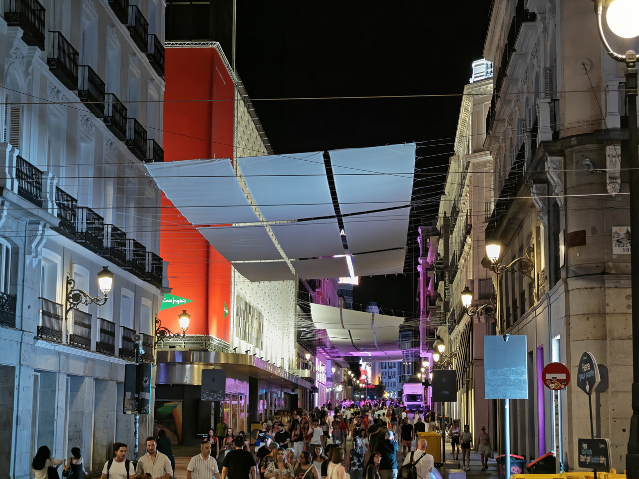 Calle iluminada de noche en Madrid con tiendas y personas paseando bajo toldos decorativos. Ambiente vibrante y urbano.