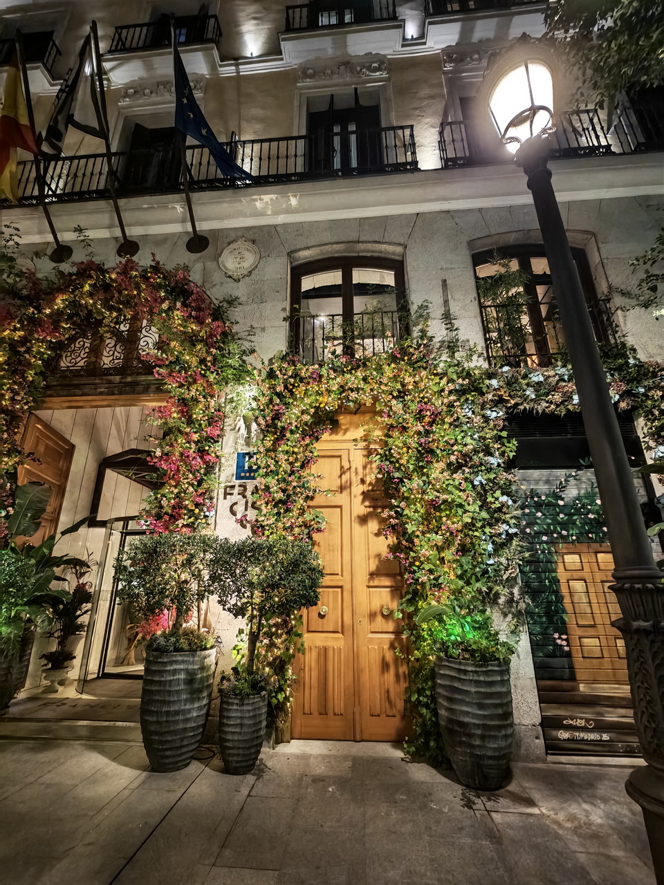 Fachada nocturna iluminada con puerta de madera, plantas trepadoras y ventanas decorativas en un edificio urbano.