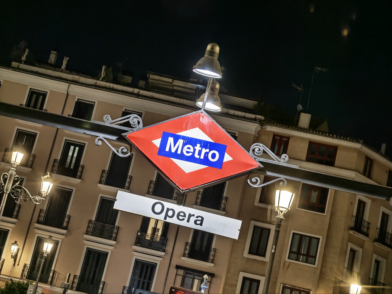 Señal de entrada a la estación de metro Ópera en Madrid, iluminada por faroles nocturnos y rodeada de edificios.