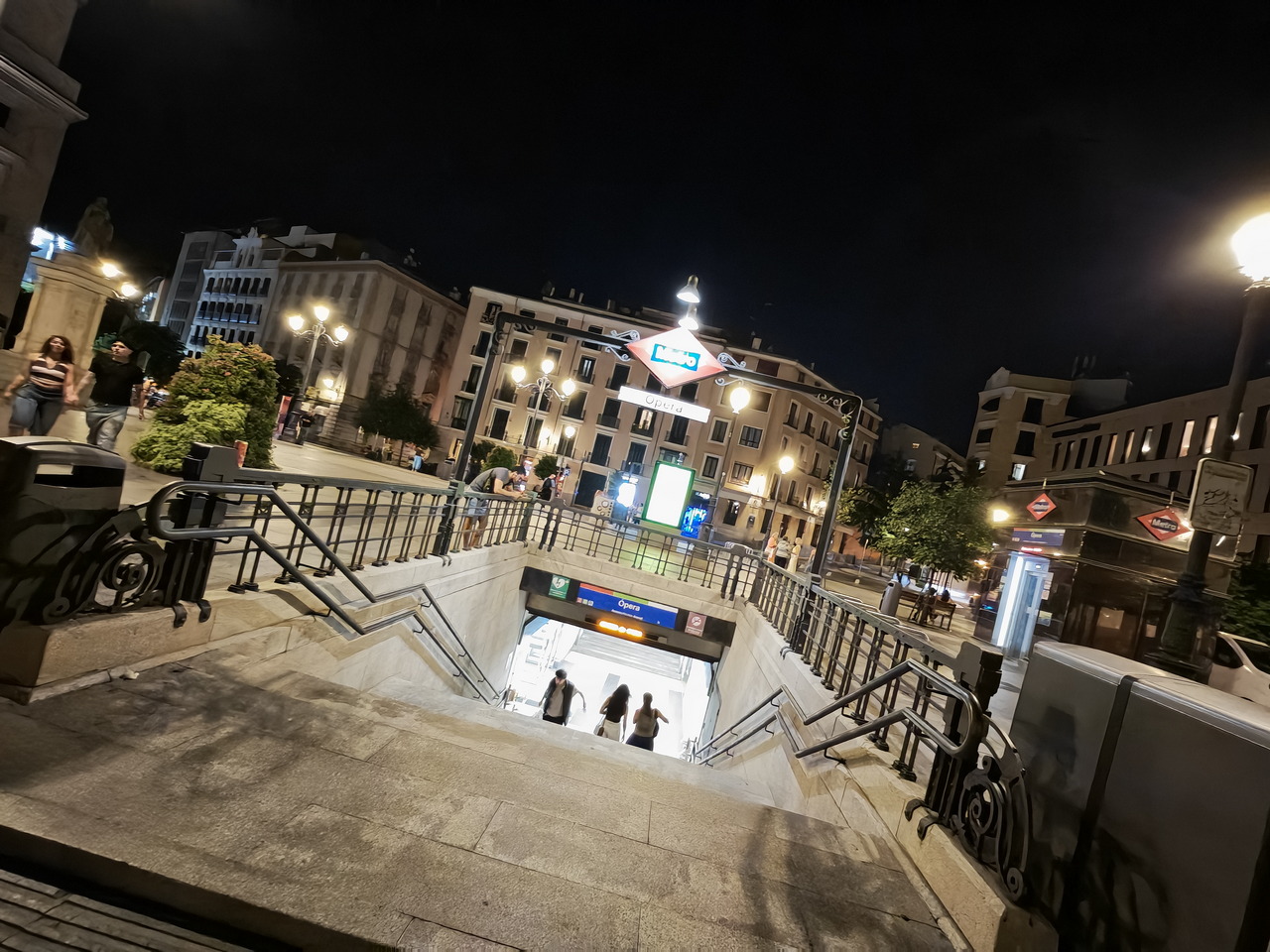 Entrada iluminada del metro Ópera en Madrid por la noche. Ambiente urbano animado con luces y transeúntes.