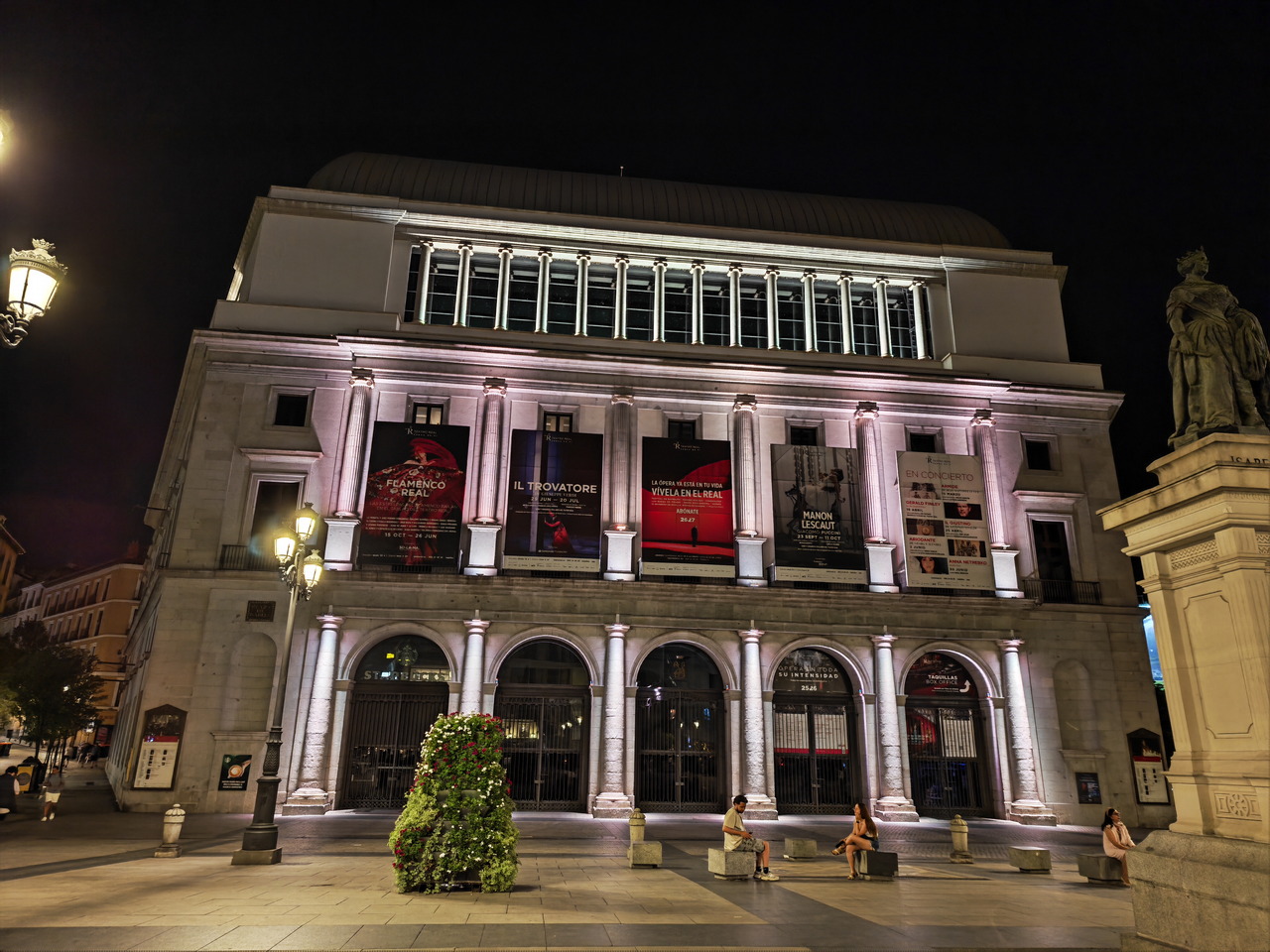 Fachada del Teatro Real de Madrid iluminada por la noche, con carteles de ópera y pocas personas en la plaza.