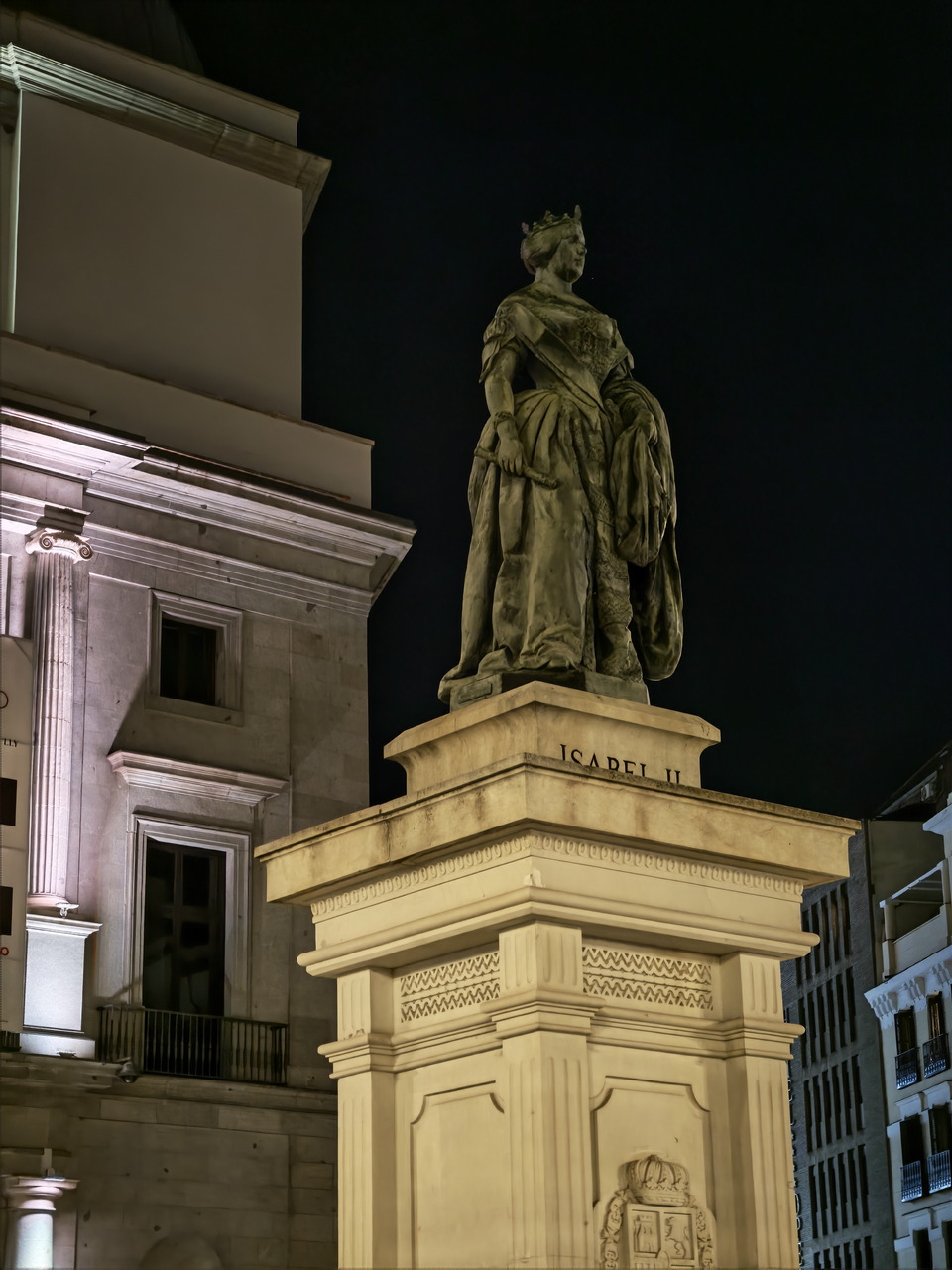 Estatua de Isabel II iluminada de noche frente al Teatro Real en Madrid, destacando su arquitectura histórica.
