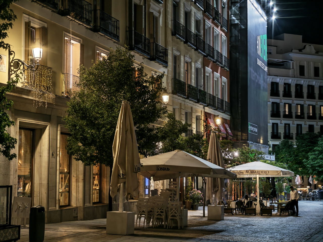 Calle nocturna en ciudad europea, con terrazas iluminadas, mesas al aire libre y edificios históricos.