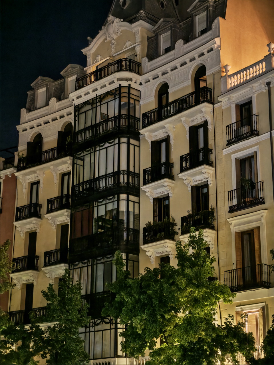 Edificio histórico iluminado de noche con balcones y árboles en la fachada.