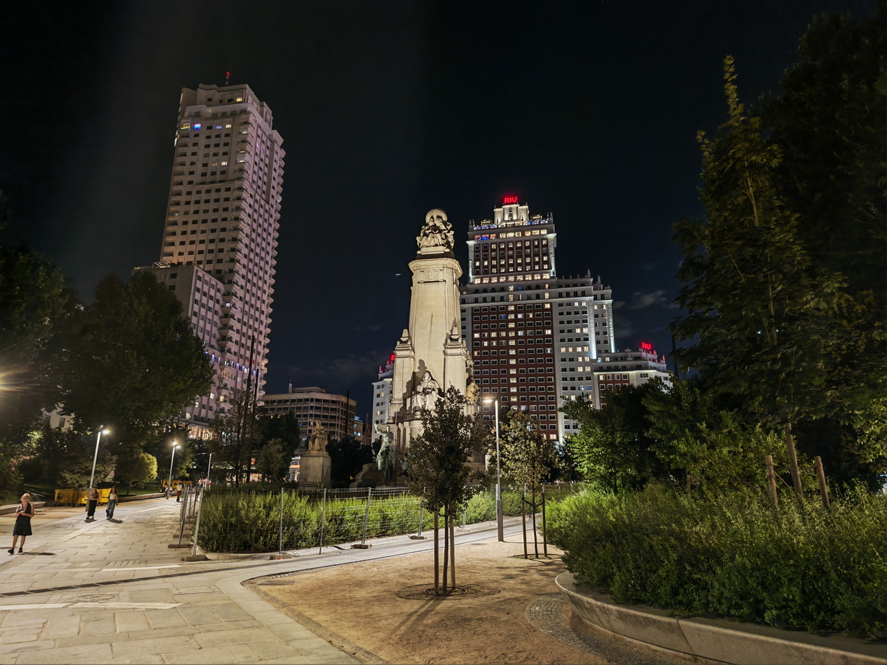 Vista nocturna de la Plaza de España, Madrid, con monumento y edificios iluminados, rodeados de árboles y jardines.