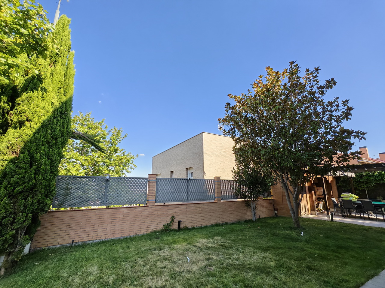 Jardín verde con árboles y seto, cercado por muro de ladrillo; cielo azul claro y casa de fondo.