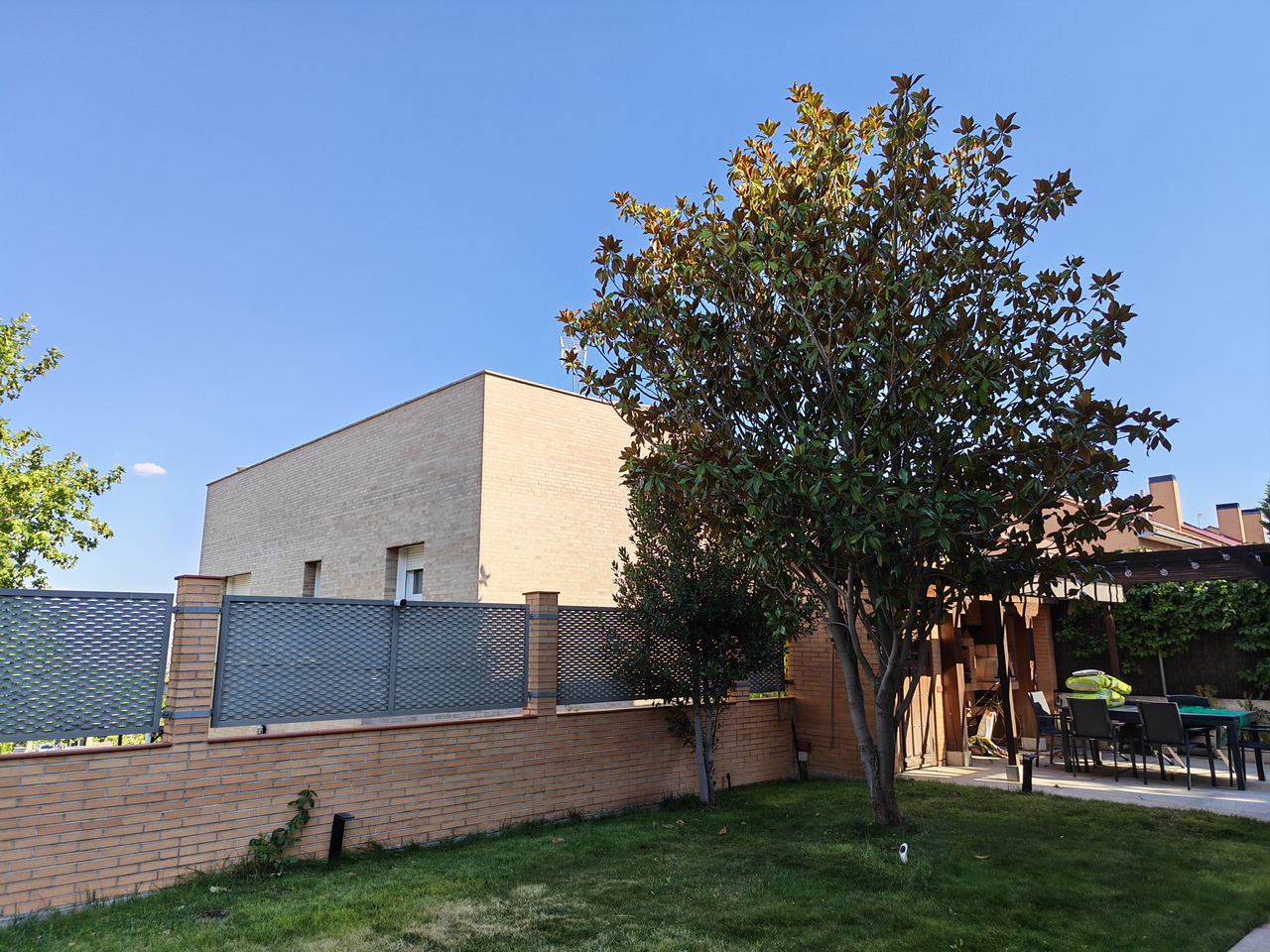 Casa moderna con jardín, seto de ladrillo y árbol bajo un cielo azul despejado. Ideal para inspiración en diseño exterior.