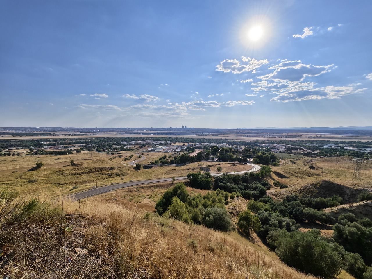Paisaje soleado con colinas doradas, carretera sinuosa, cielo azul y horizonte urbano en la distancia.