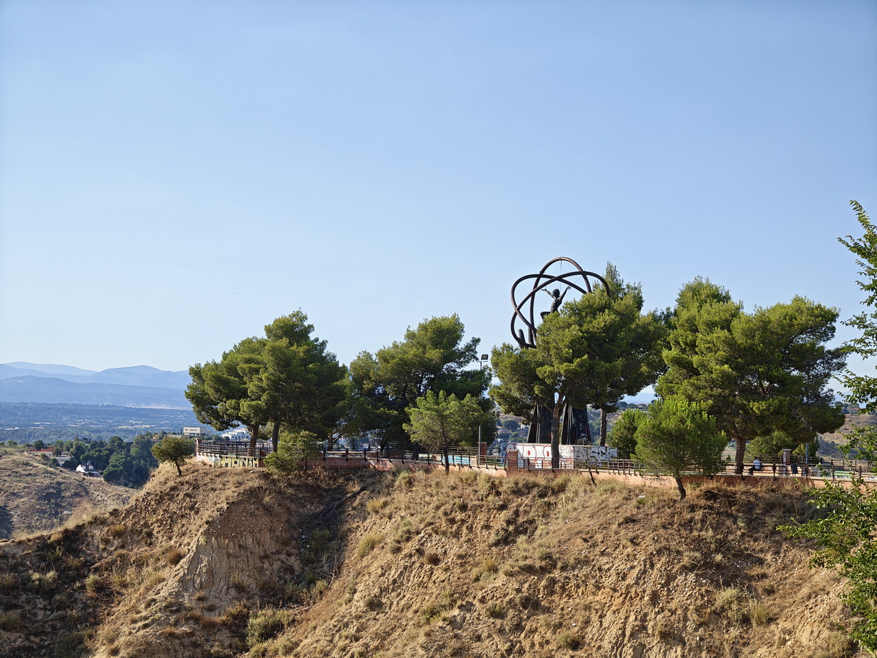 Mirador en colina con esculturas y pinos, vista panorámica de paisajes montañosos, cielo despejado.