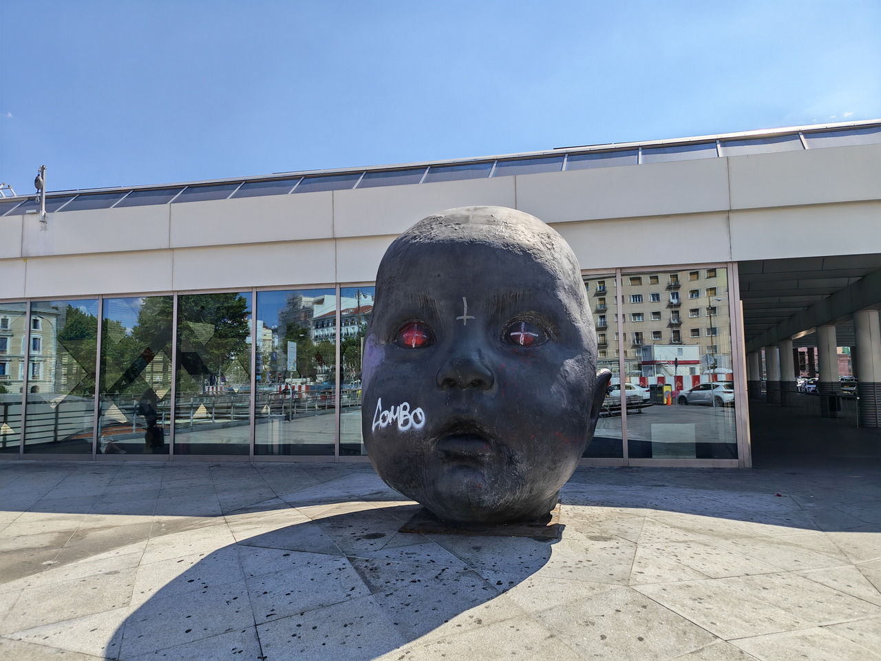 Escultura de cabeza de bebé gigante con graffiti, ubicada frente a edificio moderno con reflejos en las ventanas.