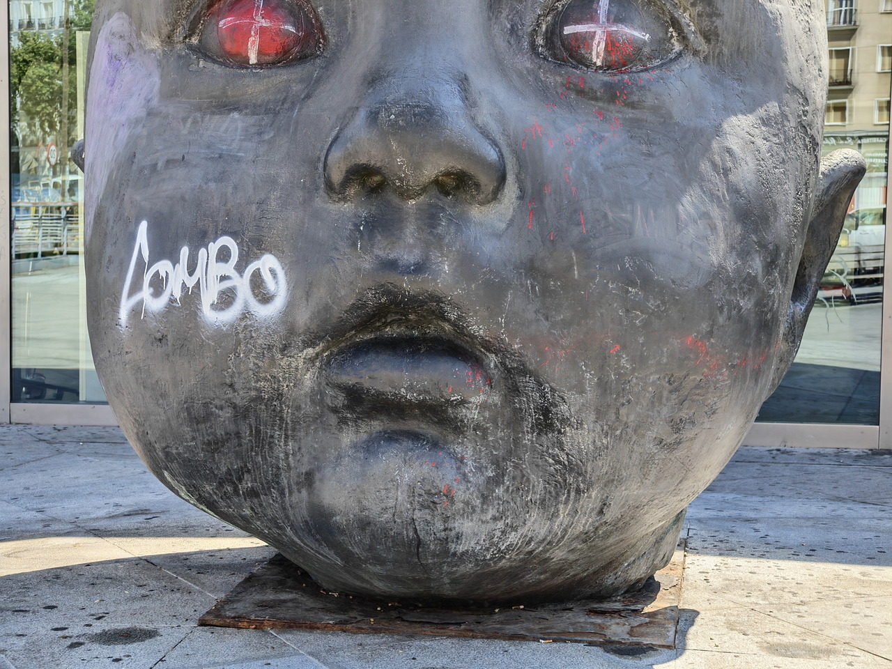 Escultura de cabeza gigante con ojos rojos y grafiti blanco en espacio urbano contemporáneo.
