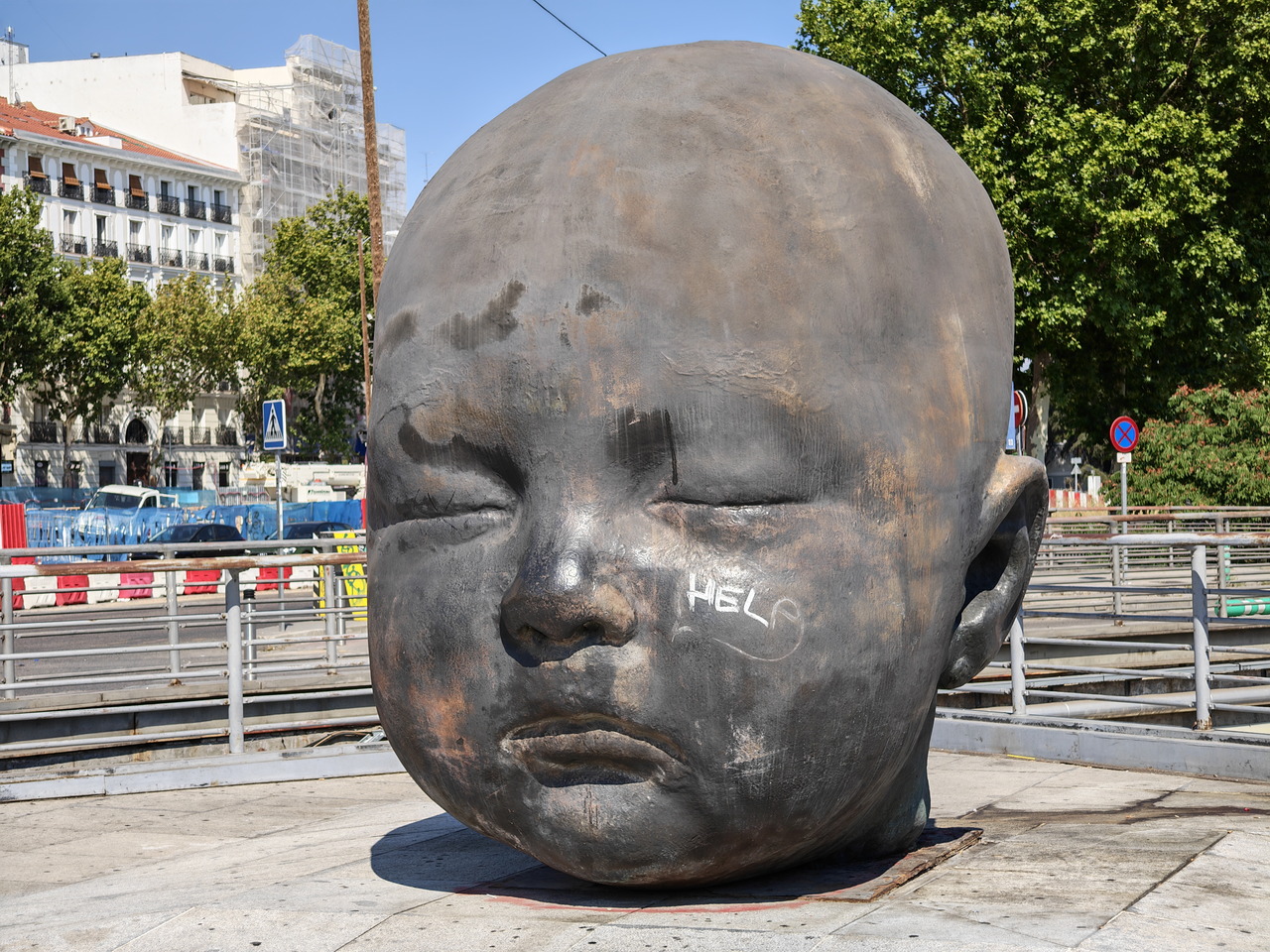 Escultura gigante de una cabeza de bebé con ojos cerrados en un espacio público, rodeada de vegetación y edificios.