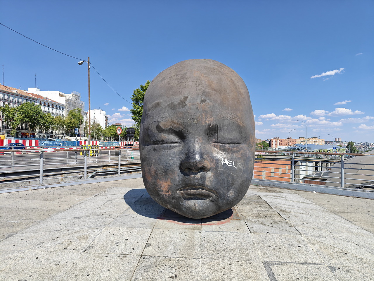 Escultura de cabeza de bebé en Plaza Colón, Madrid, con cielo despejado y edificios al fondo.